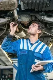 A mechanic in a blue uniform holds a flashlight up to inspect the underside of a car while holding a clipboard.