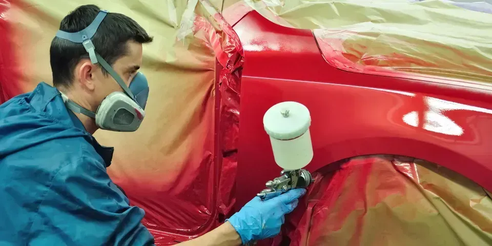 A person wearing a protective mask and gloves uses a spray gun to paint a car panel red in a workshop.