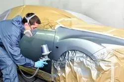 A person in a protective suit and respirator mask uses a spray gun to paint a gray car panel in an auto shop.