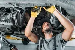 A mechanic in a grey shirt and cap works on the underside of a car, wearing yellow protective gloves.
