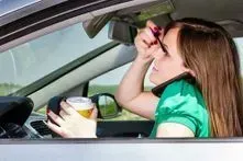 A driver in a car holds a drink with one hand while applying mascara and talking on a phone tucked against her shoulder.