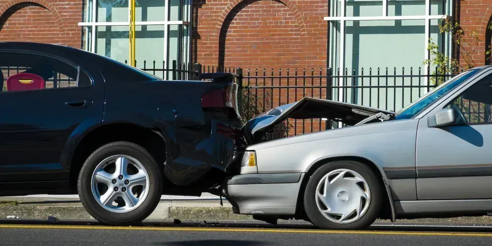 A silver car has rear-ended a dark-colored car on a street in front of a brick building.