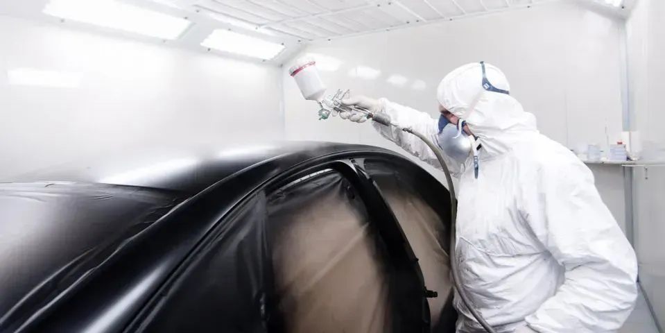 A person in protective gear uses a spray gun to apply black paint to a car in a professional spray booth.