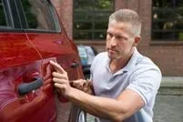 A man inspects a small dent on the side of a red car while kneeling outdoors.