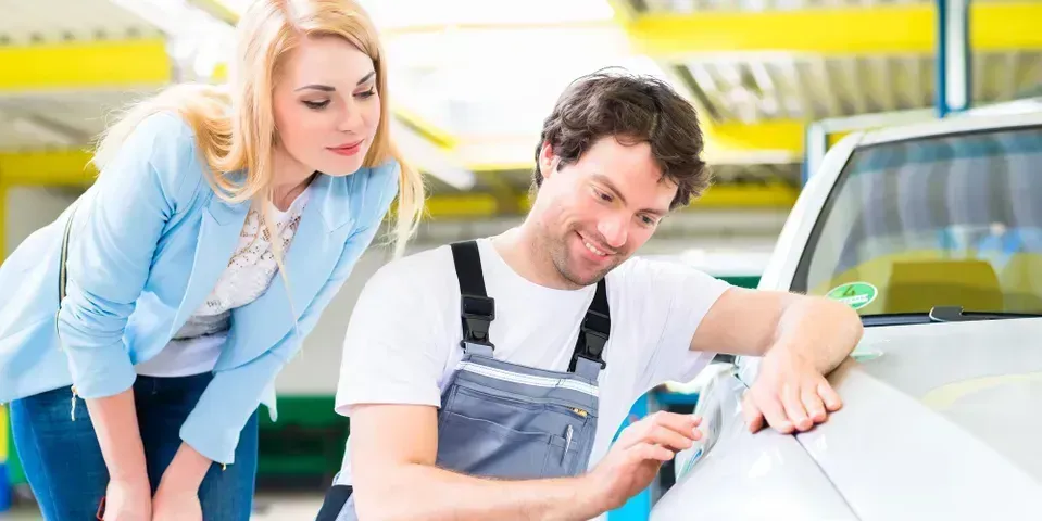 A service technician in grey overalls pointing at a scratch on a silver car while explaining it to a customer.
