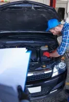 A black car in a repair shop with its front bumper removed, revealing the engine components and fluid reservoirs.