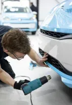 A person in a blue shirt kneels to mask a gray car in a workshop, using green paper and yellow tape.