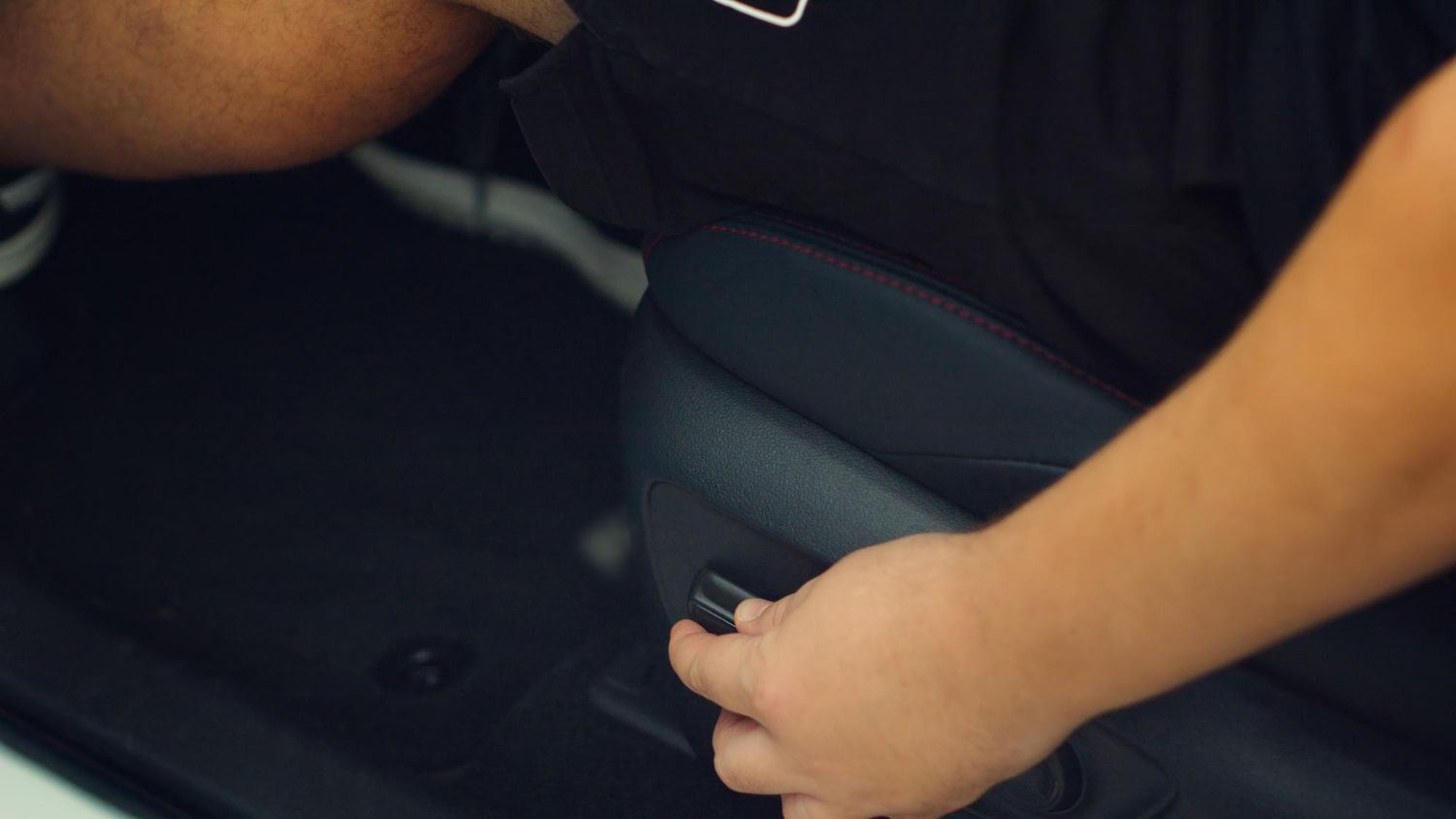 A person's hand adjusts the power seat control button on the side of a black car seat.