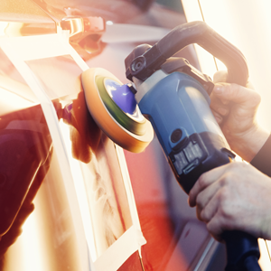 Hands using a blue electric polisher with an orange foam pad to buff a red car door surface taped off for protection.