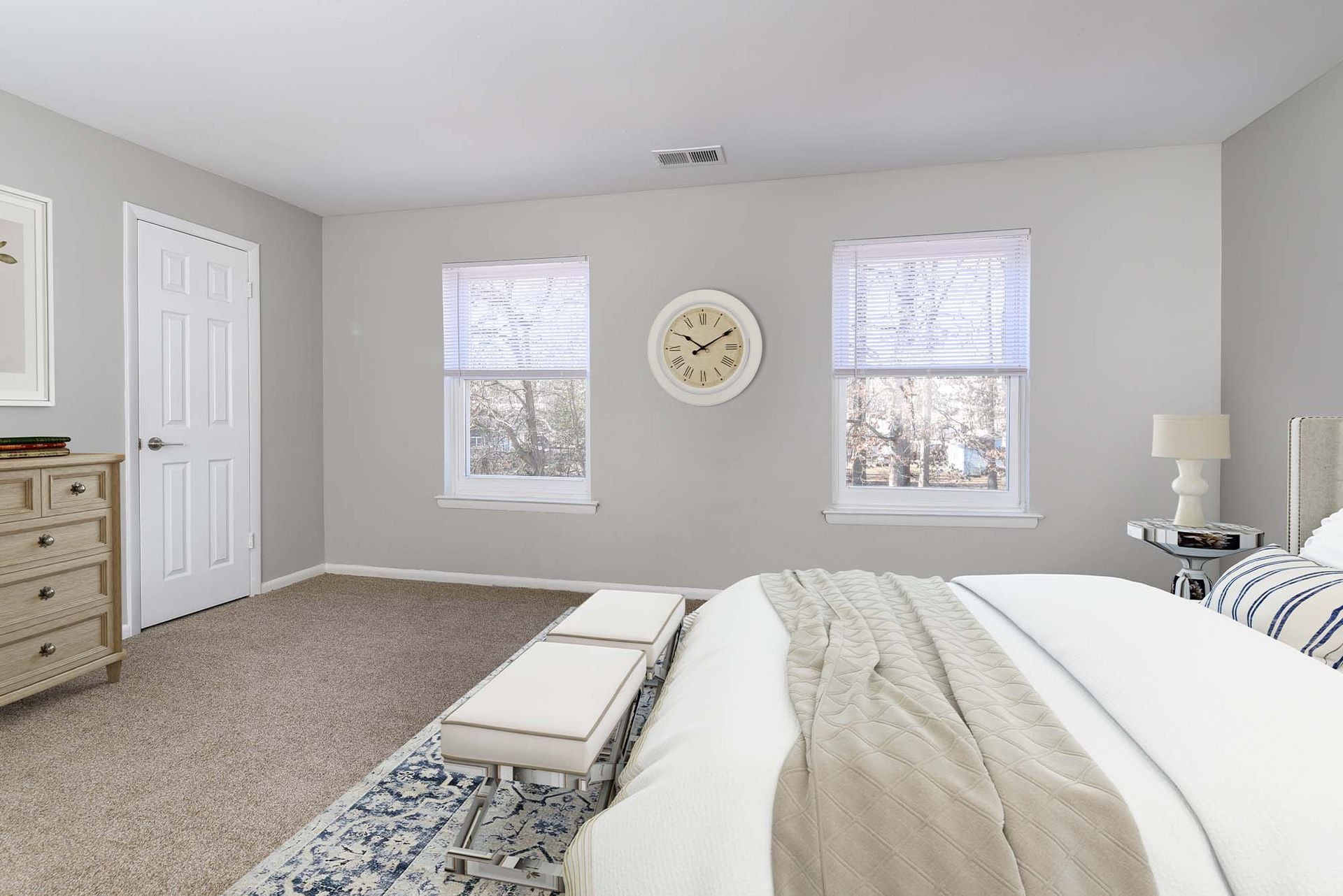 Bedroom in an apartment with a bed, bench seating, dresser, two windows, and a wall clock.