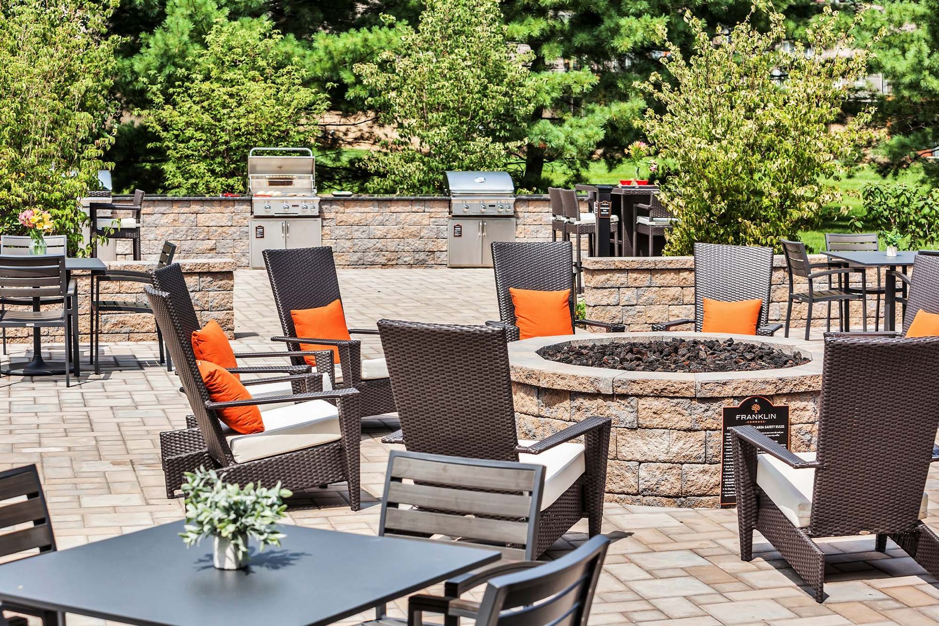 Outdoor community patio with wicker chairs around a circular stone fire pit and built-in grills.
