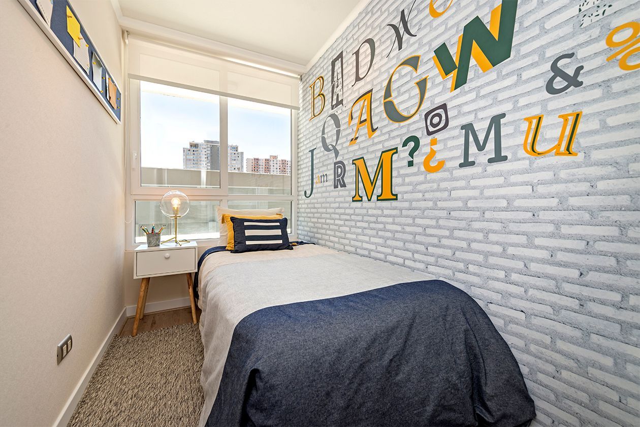 Habitación pequeña con cama, escritorio junto a la ventana y una pared decorativa de ladrillo con grandes letras de colores.