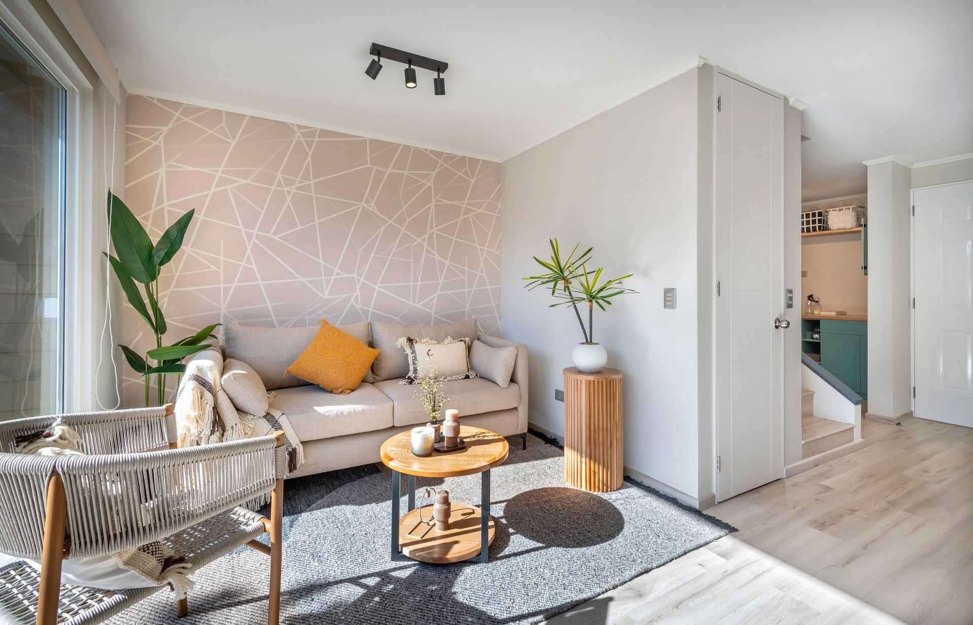 Sala de estar luminosa con sofá beige, pared decorativa estampada, plantas y una mesa de centro redonda cerca de una puerta.