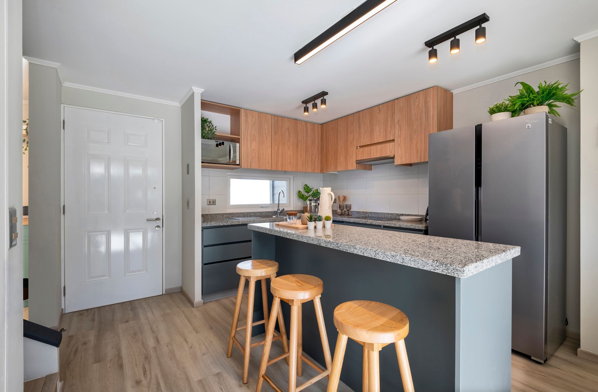 Cocina moderna con isla, encimeras de granito, armarios de madera, nevera de acero inoxidable y tres taburetes.