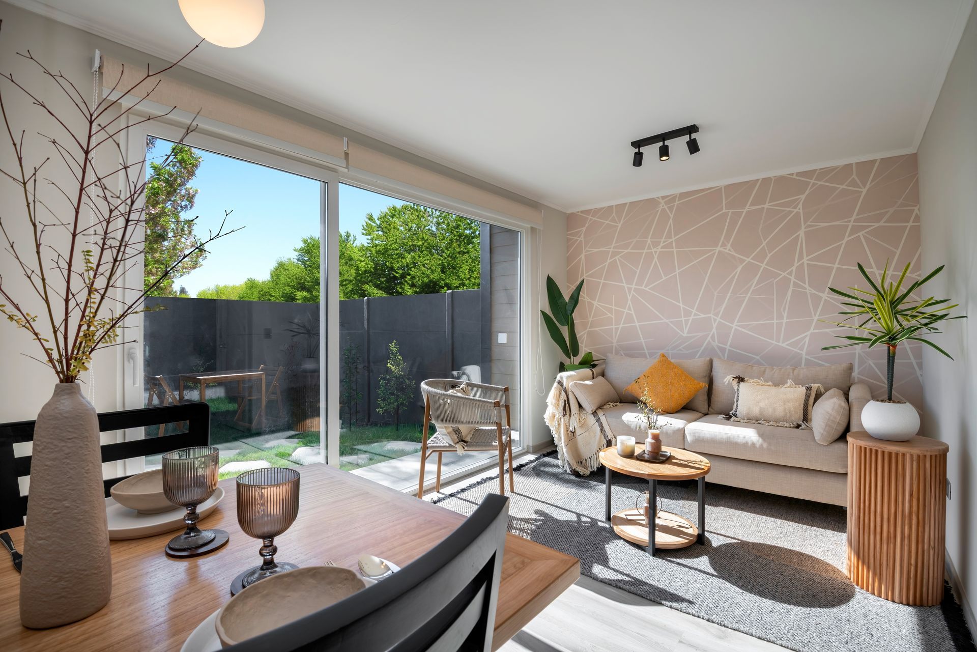 Sala de estar moderna y luminosa con sofá beige, puertas de cristal, mesa de comedor de madera y plantas en macetas.