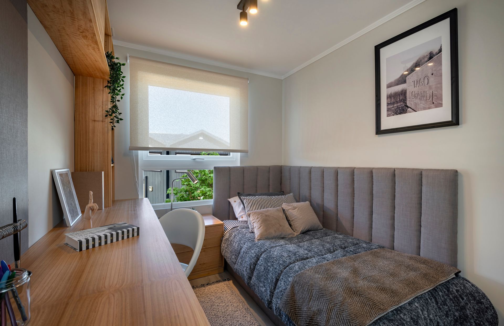 Habitación pequeña con cama gris, escritorio de madera, ventana y cuadro enmarcado.