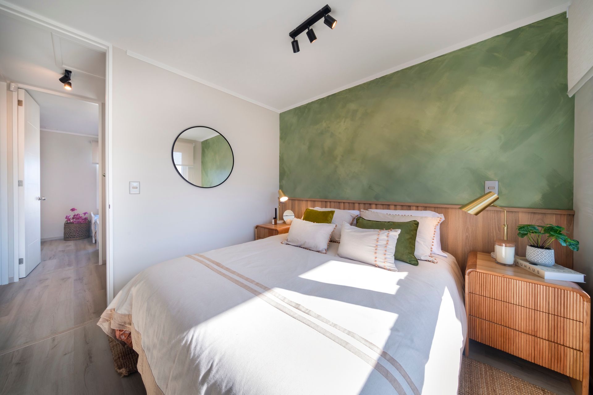 Habitación luminosa con una pared decorativa verde, cabecero de madera, espejo redondo y luz natural sobre la cama.