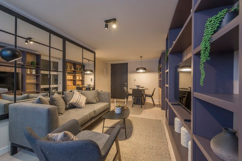 Sala de estar moderna con sofá gris, mampara de cristal, estanterías empotradas e iluminación cálida.