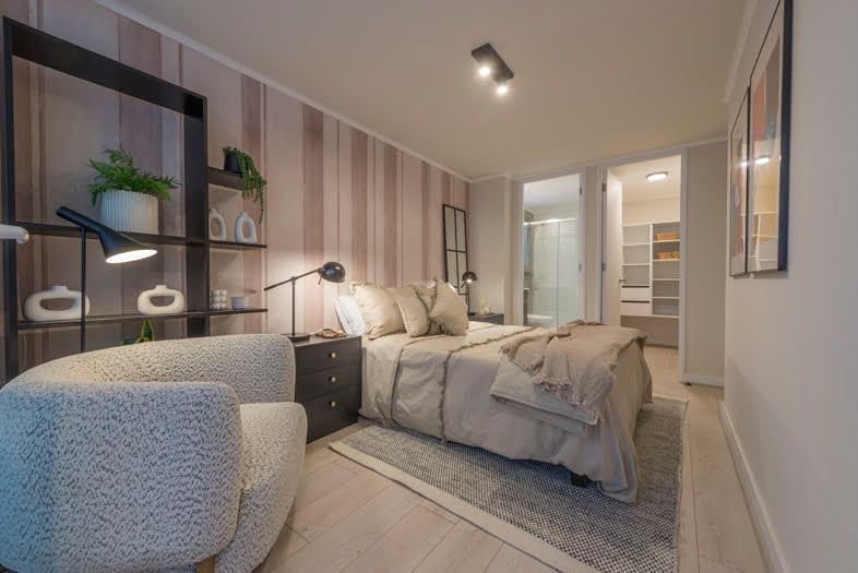 Acogedor dormitorio moderno con ropa de cama beige, silla gris, estanterías negras y lámparas colgantes de luz tenue.