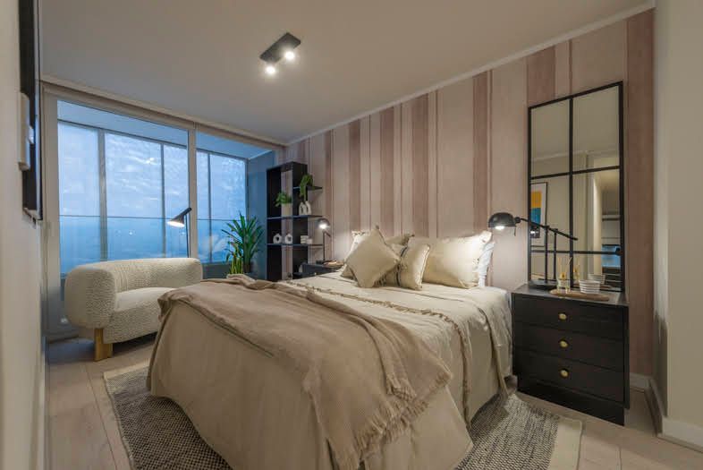 Dormitorio moderno con cama beige, pared decorativa a rayas, tocador, silla y gran ventanal con vistas a los árboles.