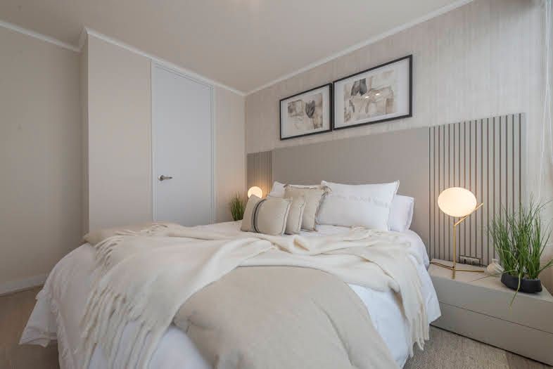 Dormitorio minimalista de color beige con cama sin hacer, lámpara de noche, cuadro enmarcado y paneles de pared a rayas.