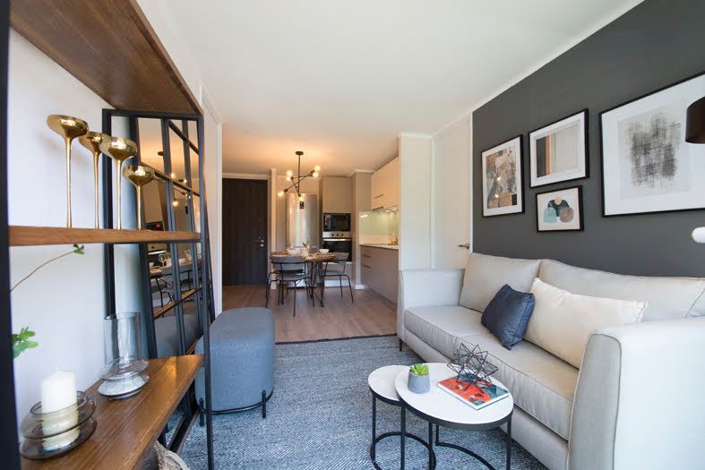 Sala de estar moderna con sofá beige, alfombra gris, cuadro en la pared y cocina visible al fondo.