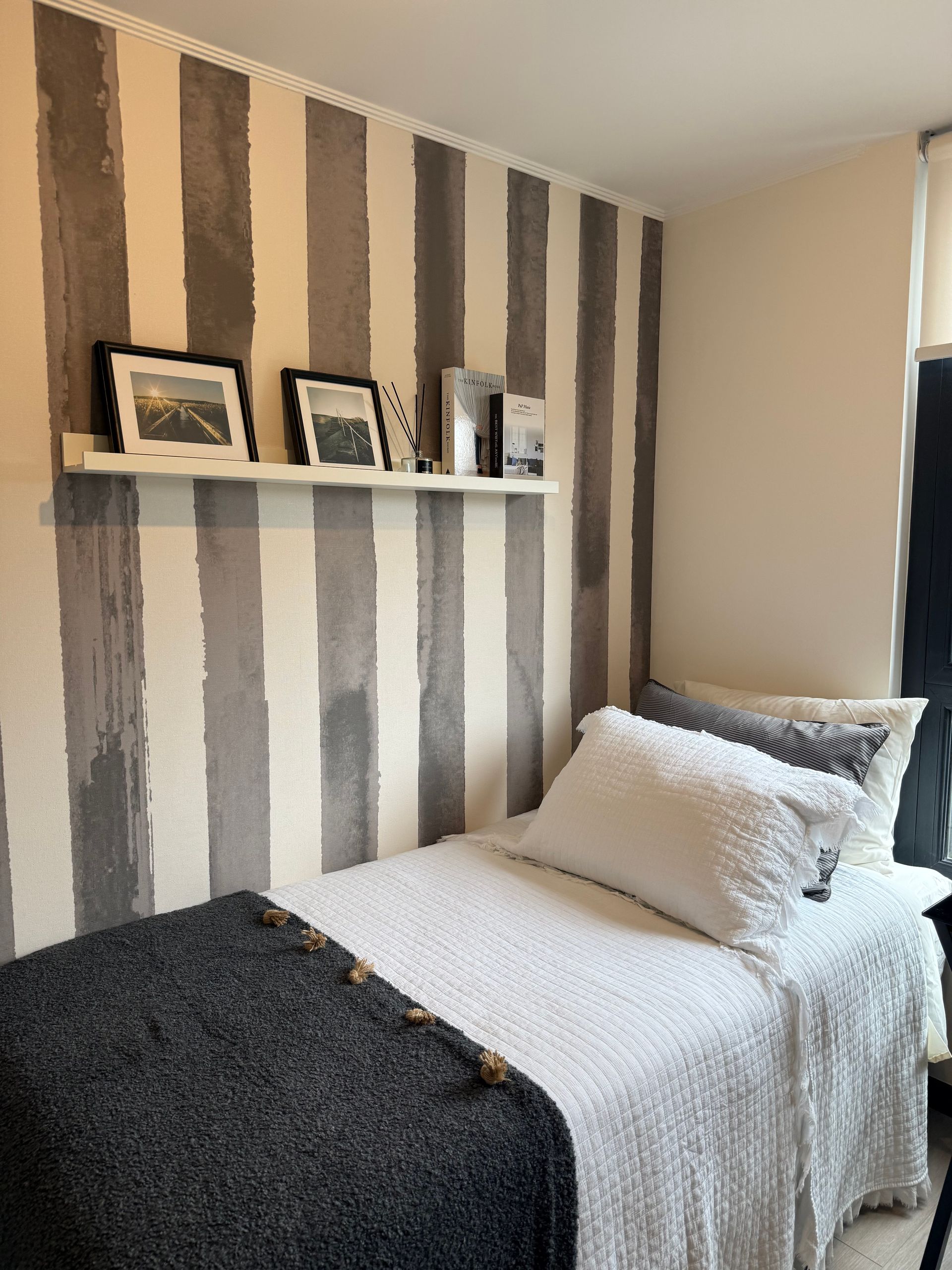 Habitación pequeña con pared decorativa a rayas, cama estrecha y estante flotante con fotos enmarcadas.