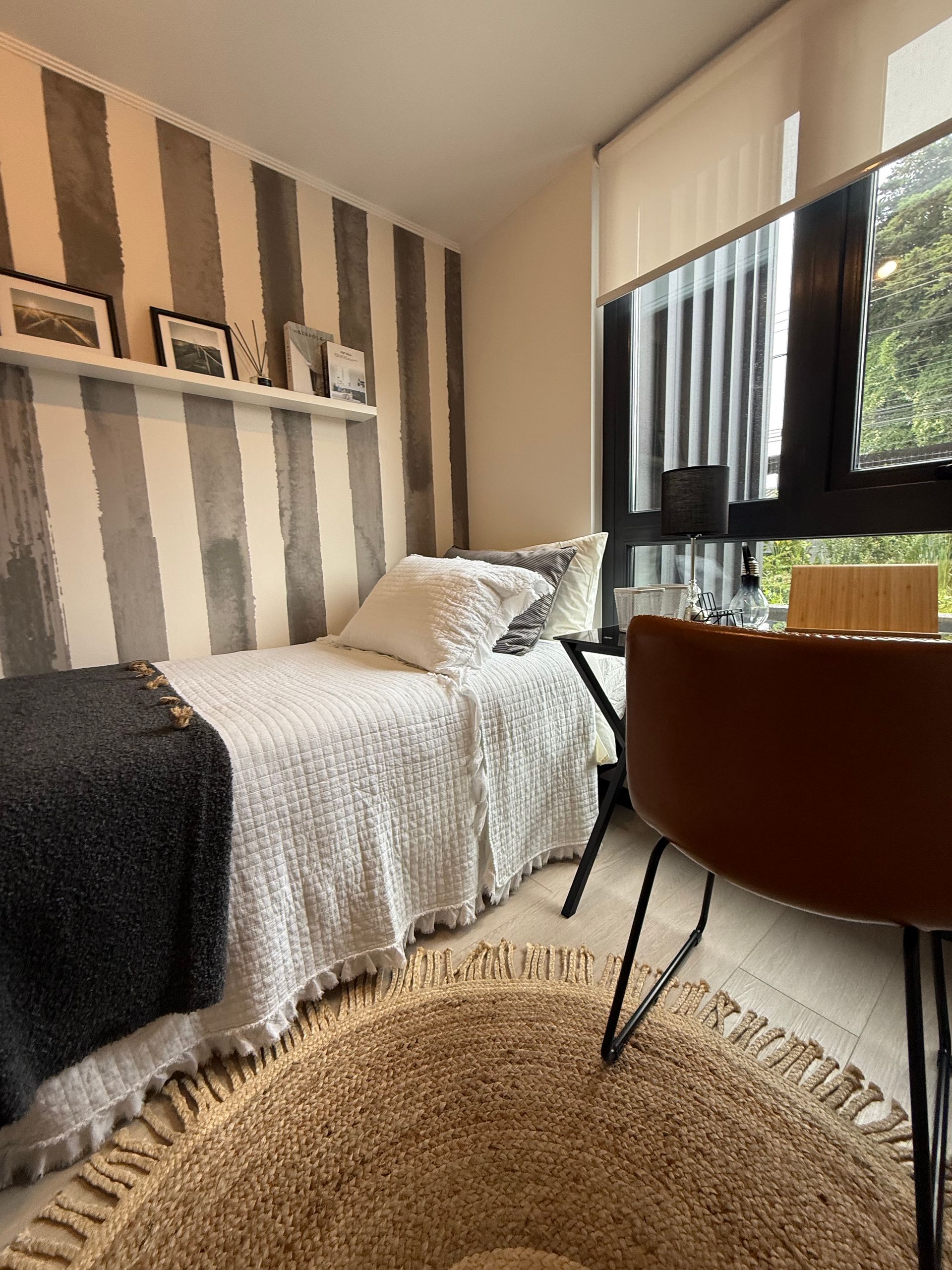 Acogedora habitación con cama, papel pintado a rayas, alfombra redonda, silla de escritorio y ventana con persianas.