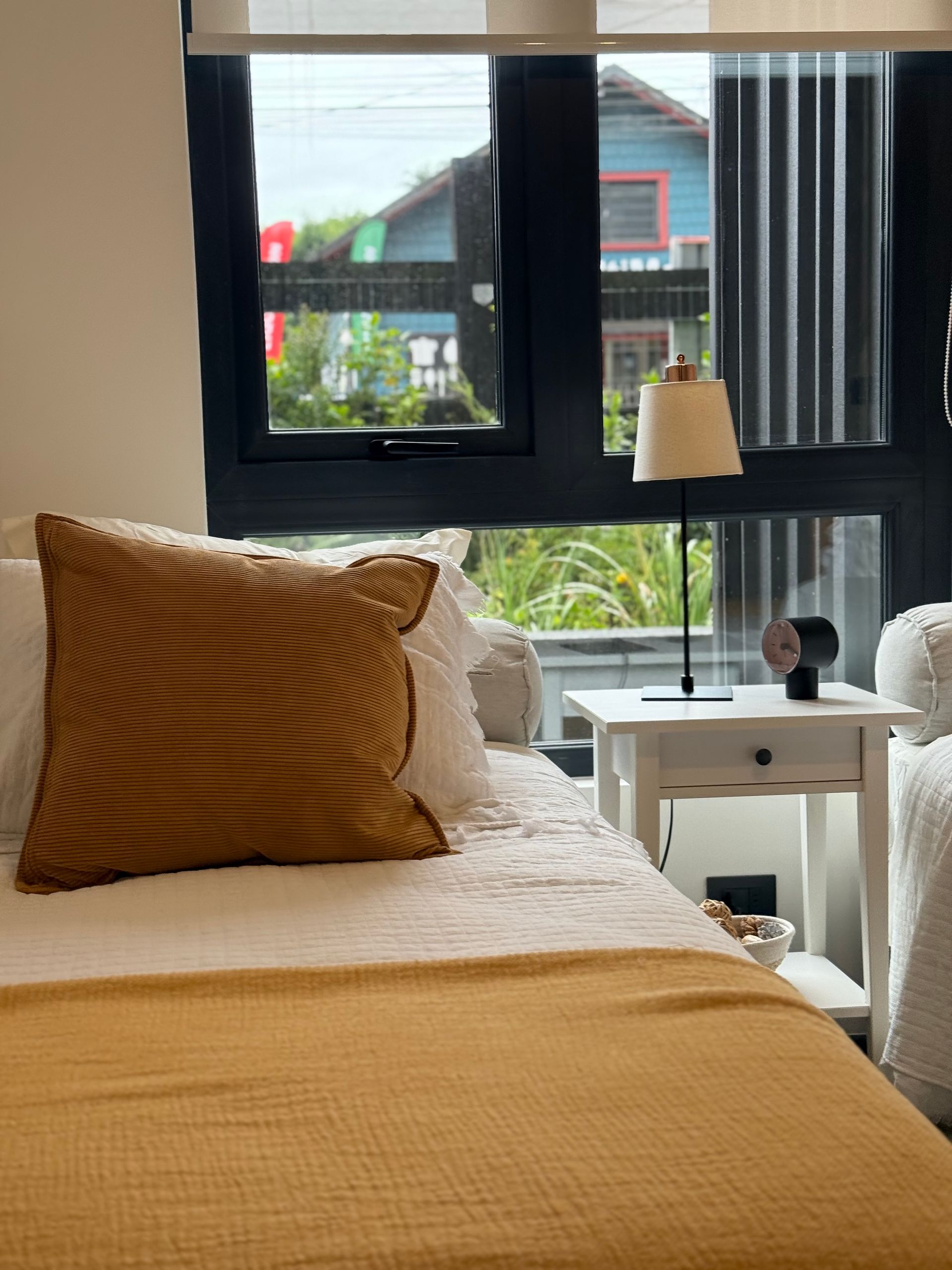 Habitación acogedora con colcha y almohada color mostaza, mesita de noche y grandes ventanales con vista a la ciudad.