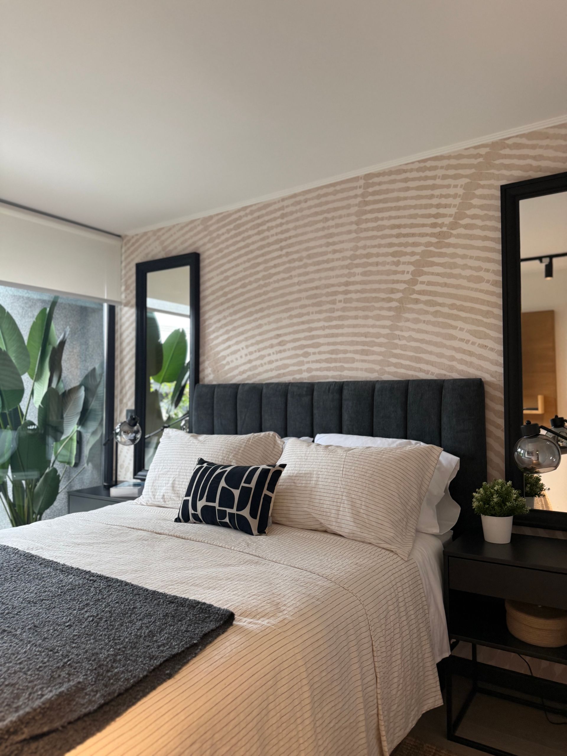 Dormitorio moderno con cama tapizada en negro, ropa de cama blanca, almohada estampada y un gran espejo junto a plantas.