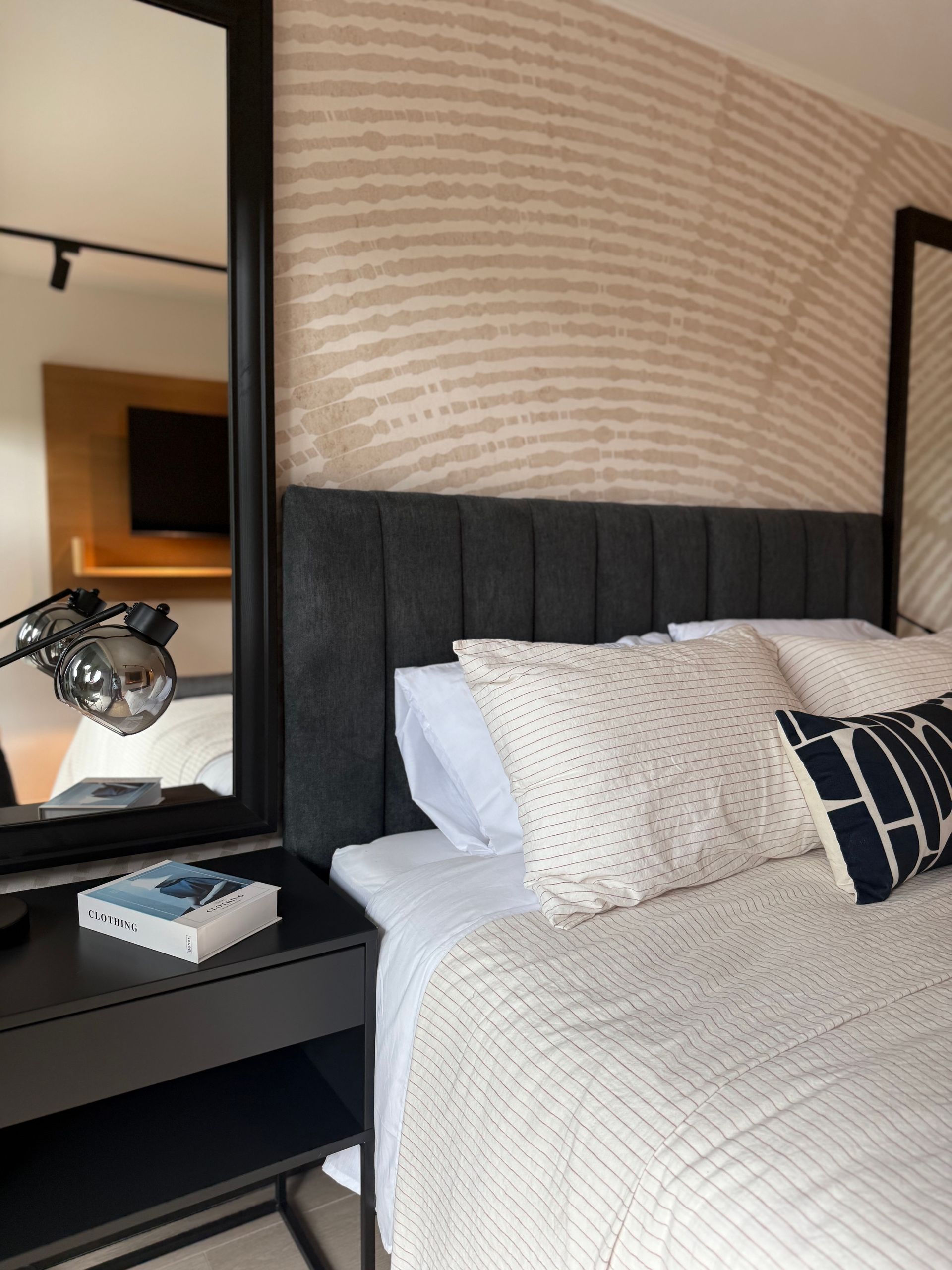 Dormitorio moderno con cabecero capitoné negro, cojines estampados y mesita de noche con espejo.