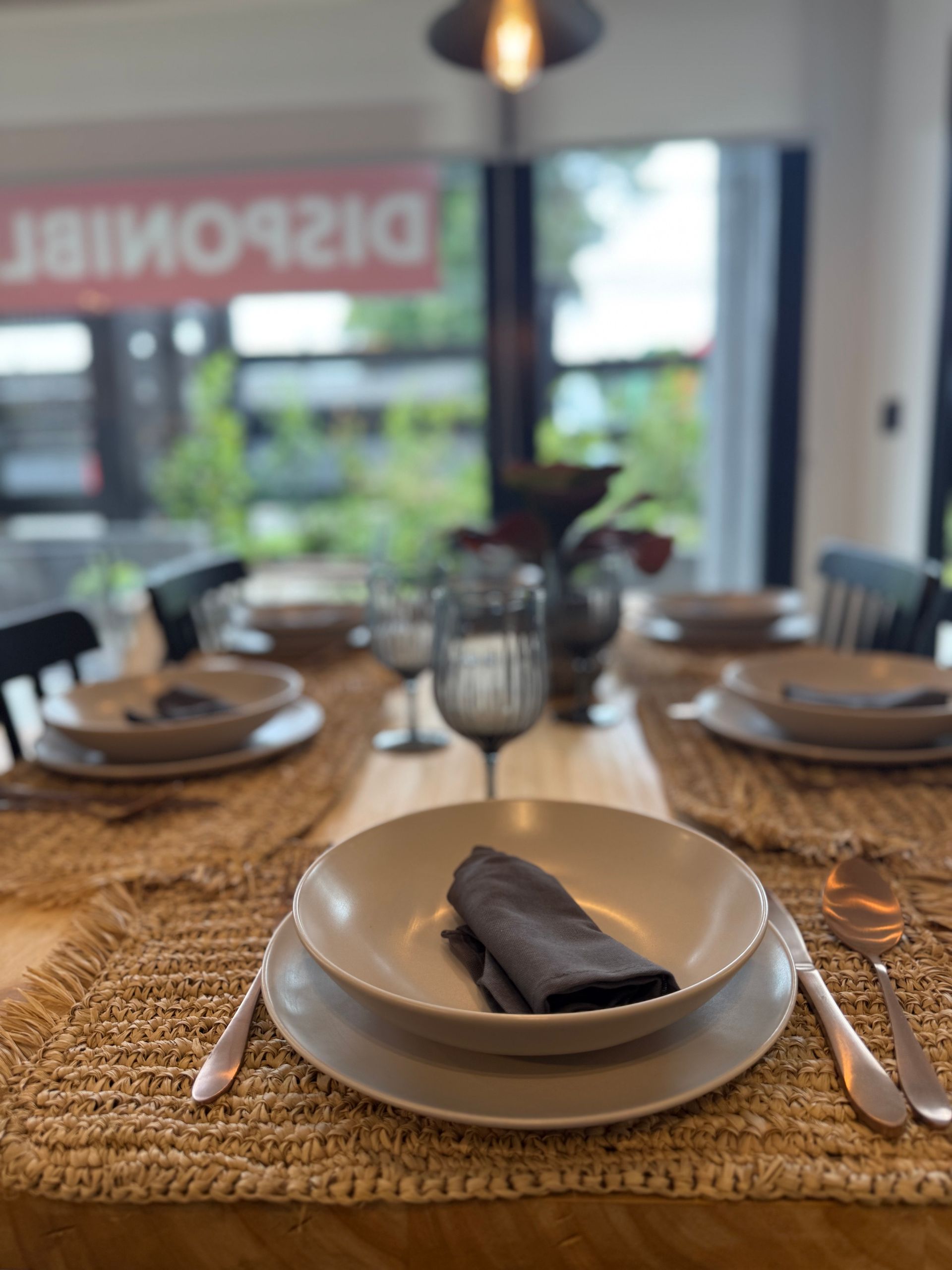 Mesa de comedor puesta con manteles individuales tejidos, platos blancos y servilletas junto a un restaurante con ventanales.