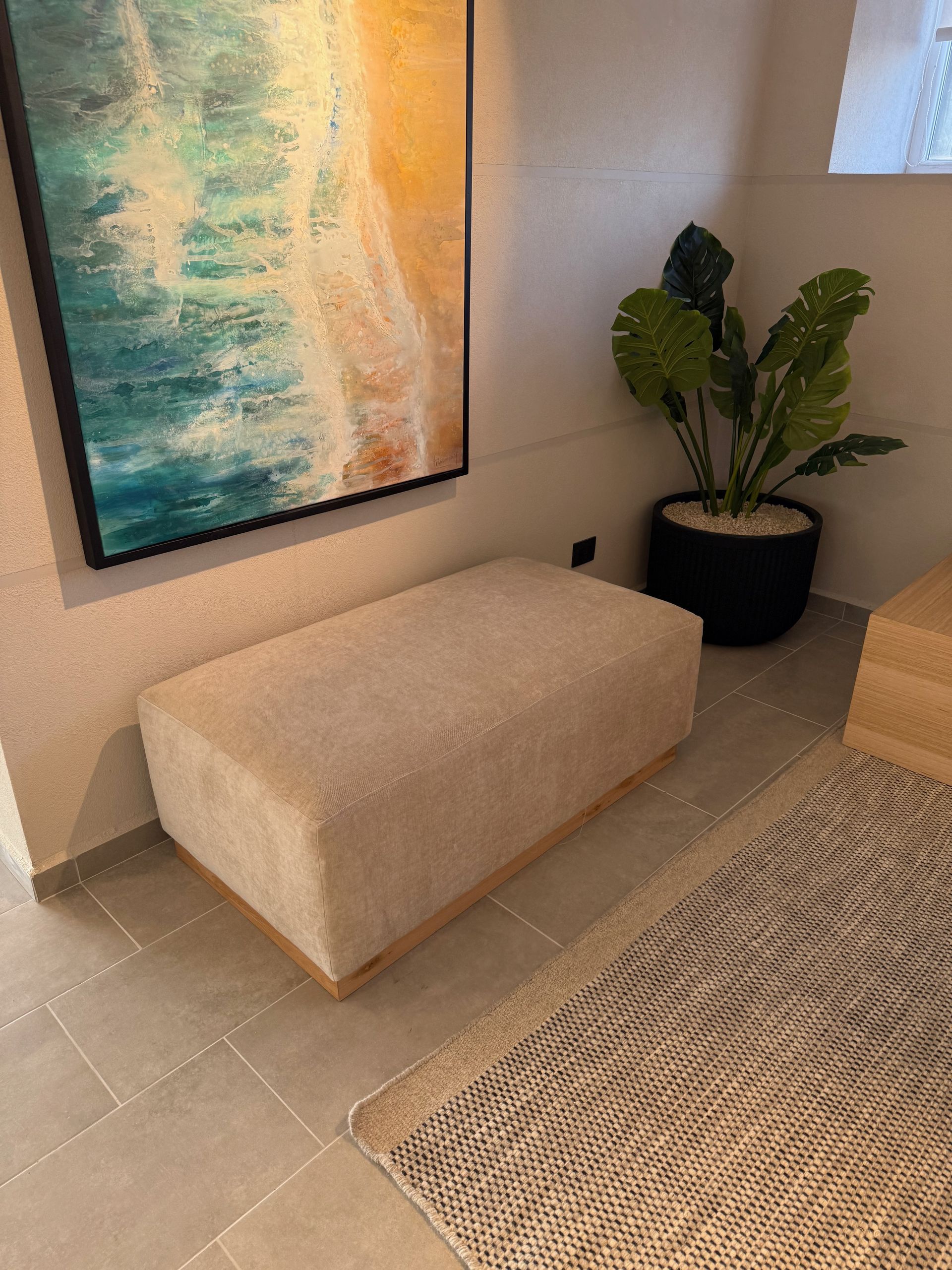 Entrada moderna con banco beige, arte abstracto en la pared, planta en maceta y alfombra tejida sobre suelo de baldosas.