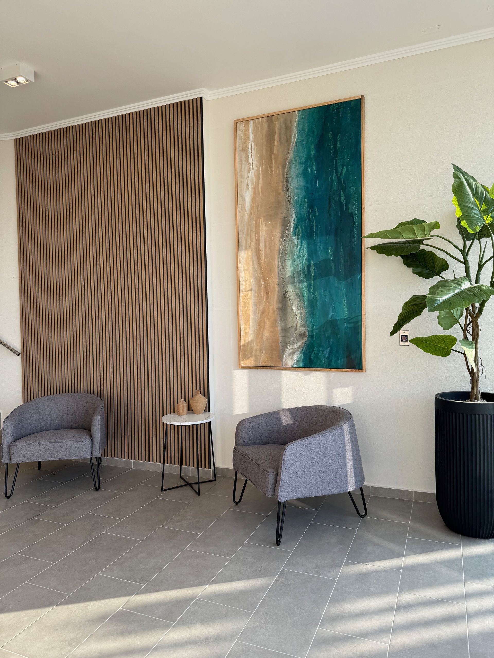 Salón moderno con dos sillas grises, arte abstracto en la pared y una gran planta en maceta, bañado por la luz del sol.