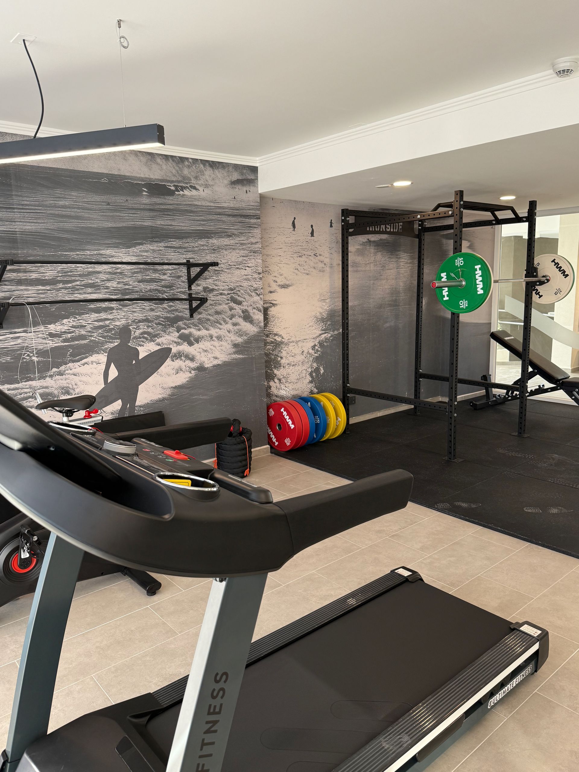 Gimnasio con cinta de correr, soporte para pesas, mancuernas y aparatos de ejercicio contra una pared gris.