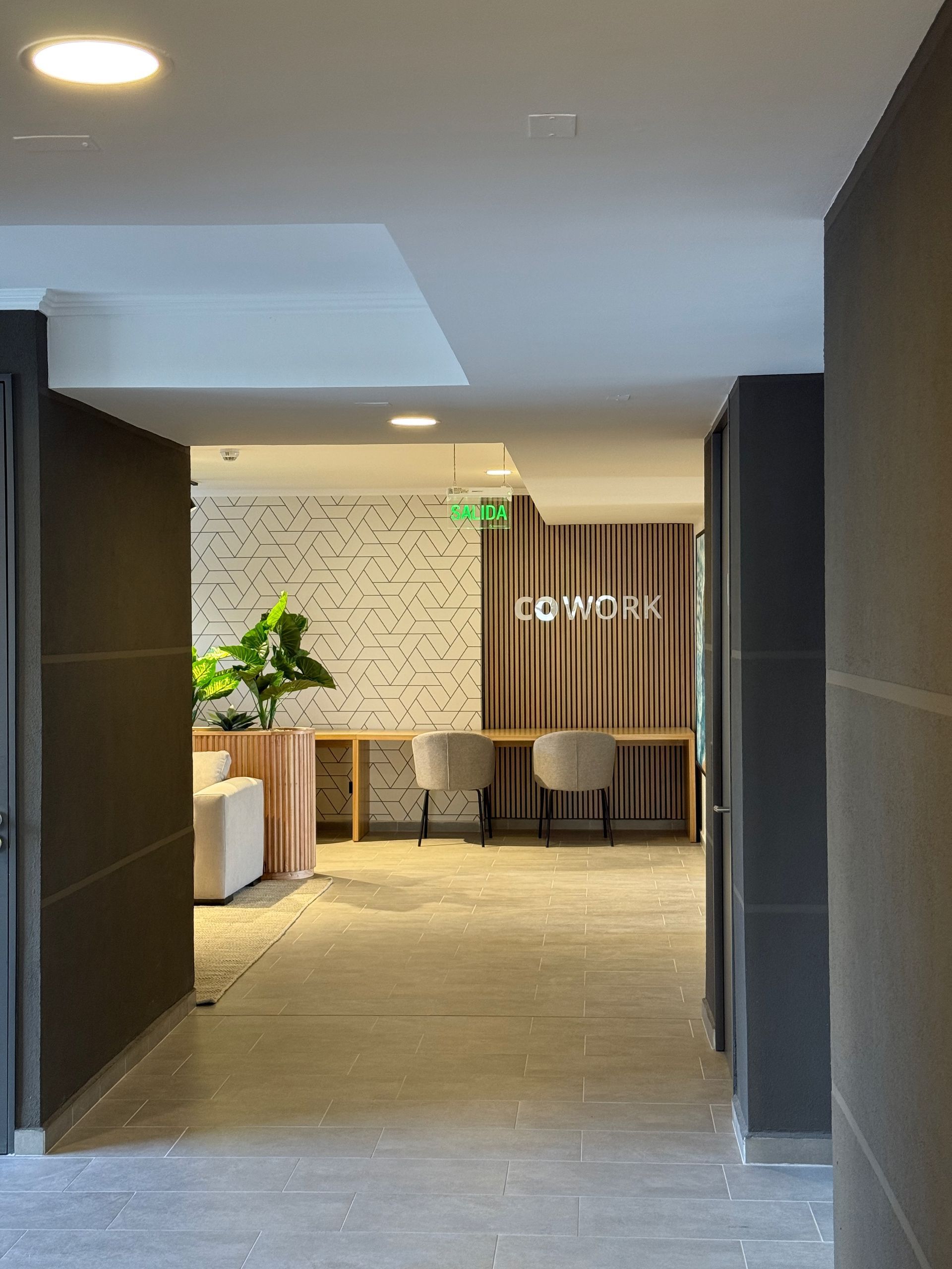 Pasillo que da paso a un salón moderno con mesas, sillas, plantas y detalles de pared revestidos con listones de madera.