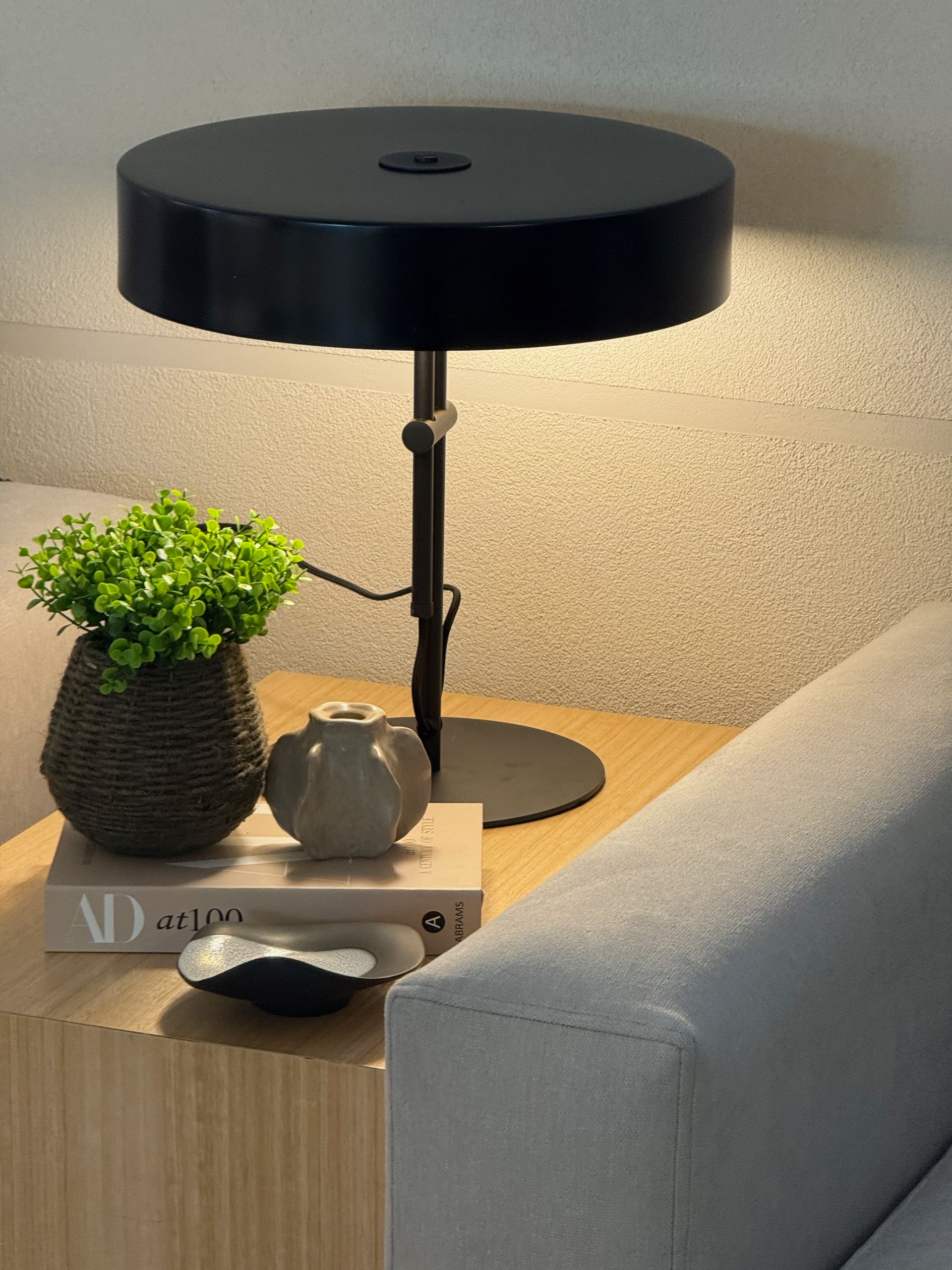 Lámpara de mesa negra sobre una mesita auxiliar junto a una pequeña planta, una tetera y un libro, al lado de un sofá gris.