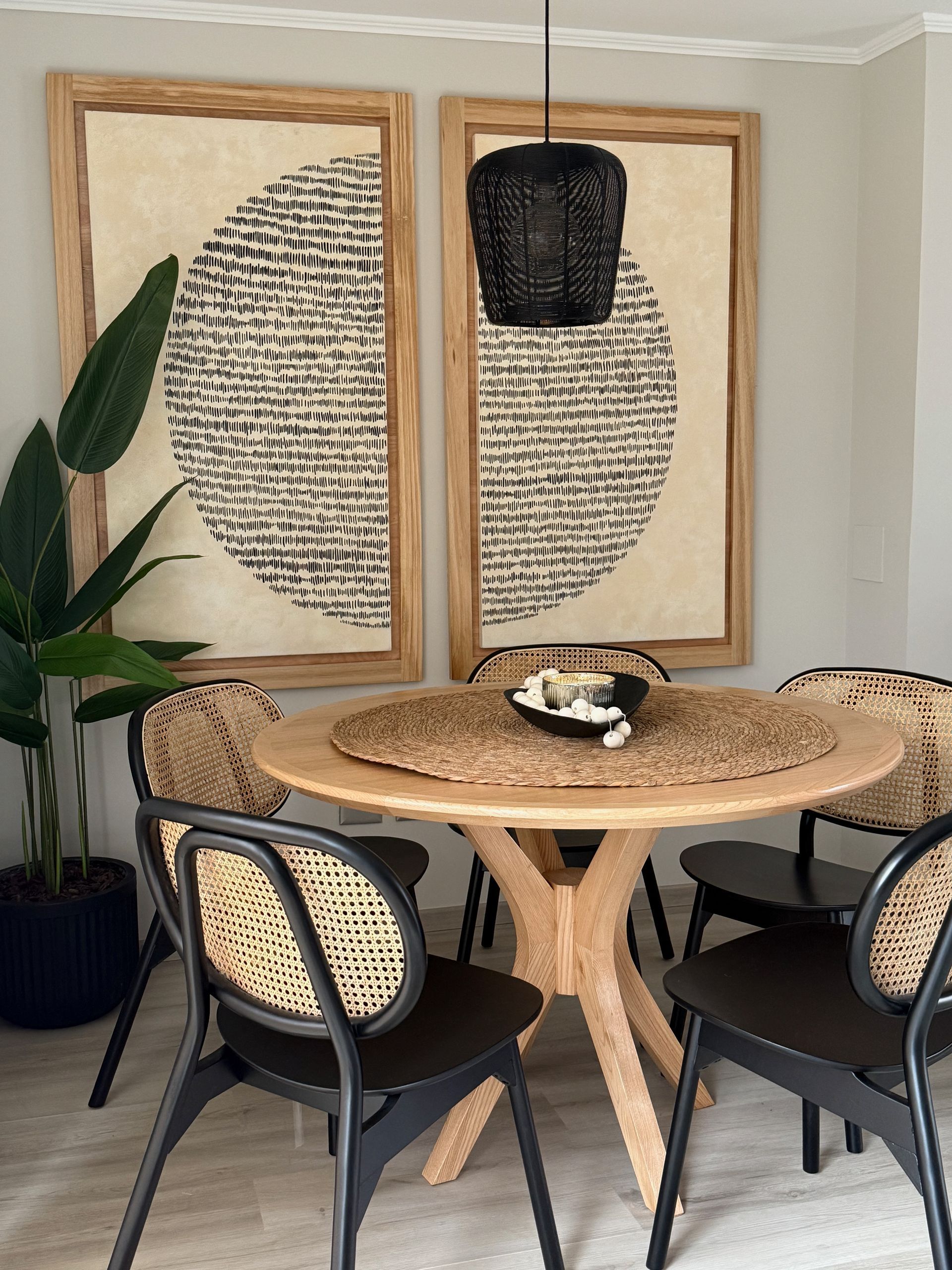 Acogedor rincón de comedor con mesa de ratán, sillas negras, una gran planta en maceta y un cuadro abstracto enmarcado.