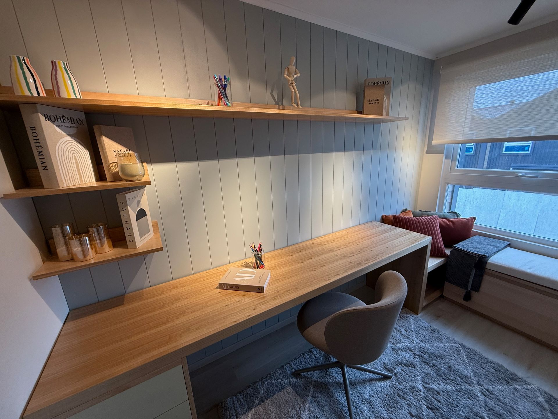 Oficina moderna en casa con escritorio de madera, silla gris, estantes de pared y un asiento junto a una ventana luminosa.