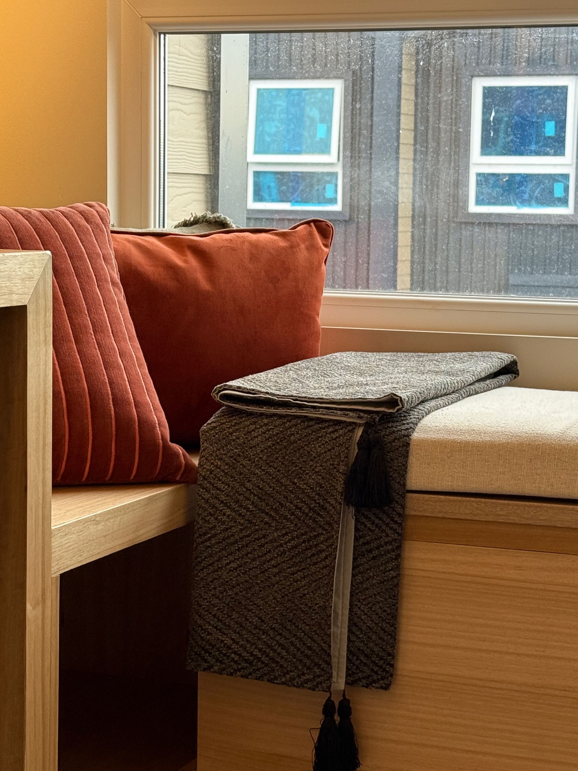 Acogedor asiento junto a la ventana con cojines color óxido y beige, una mesita auxiliar de mimbre.