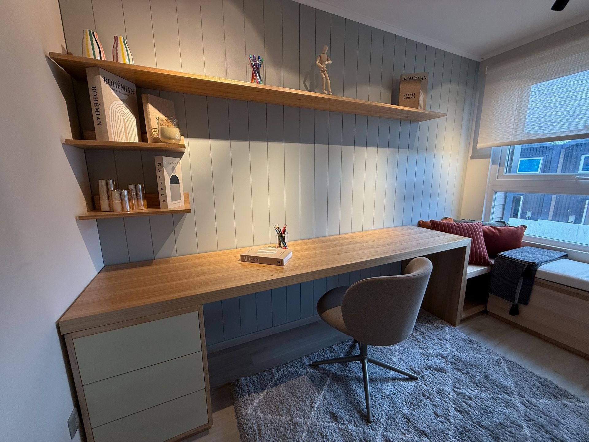 Despacho minimalista en casa con escritorio de madera, estanterías, silla gris y asiento junto a una gran ventana.