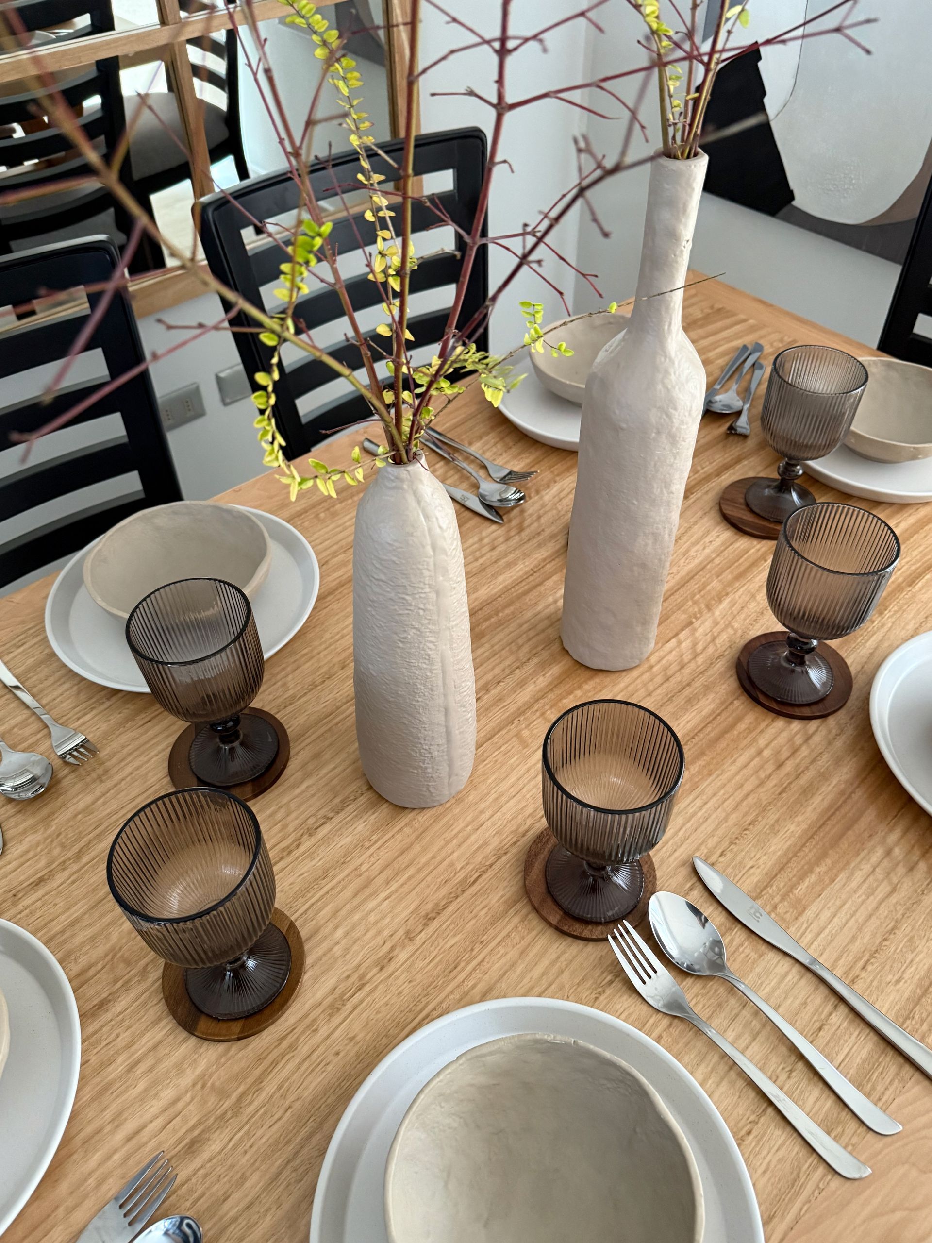 Mesa de comedor puesta con jarrones blancos, cristalería marrón, platos y cubiertos en una habitación luminosa.