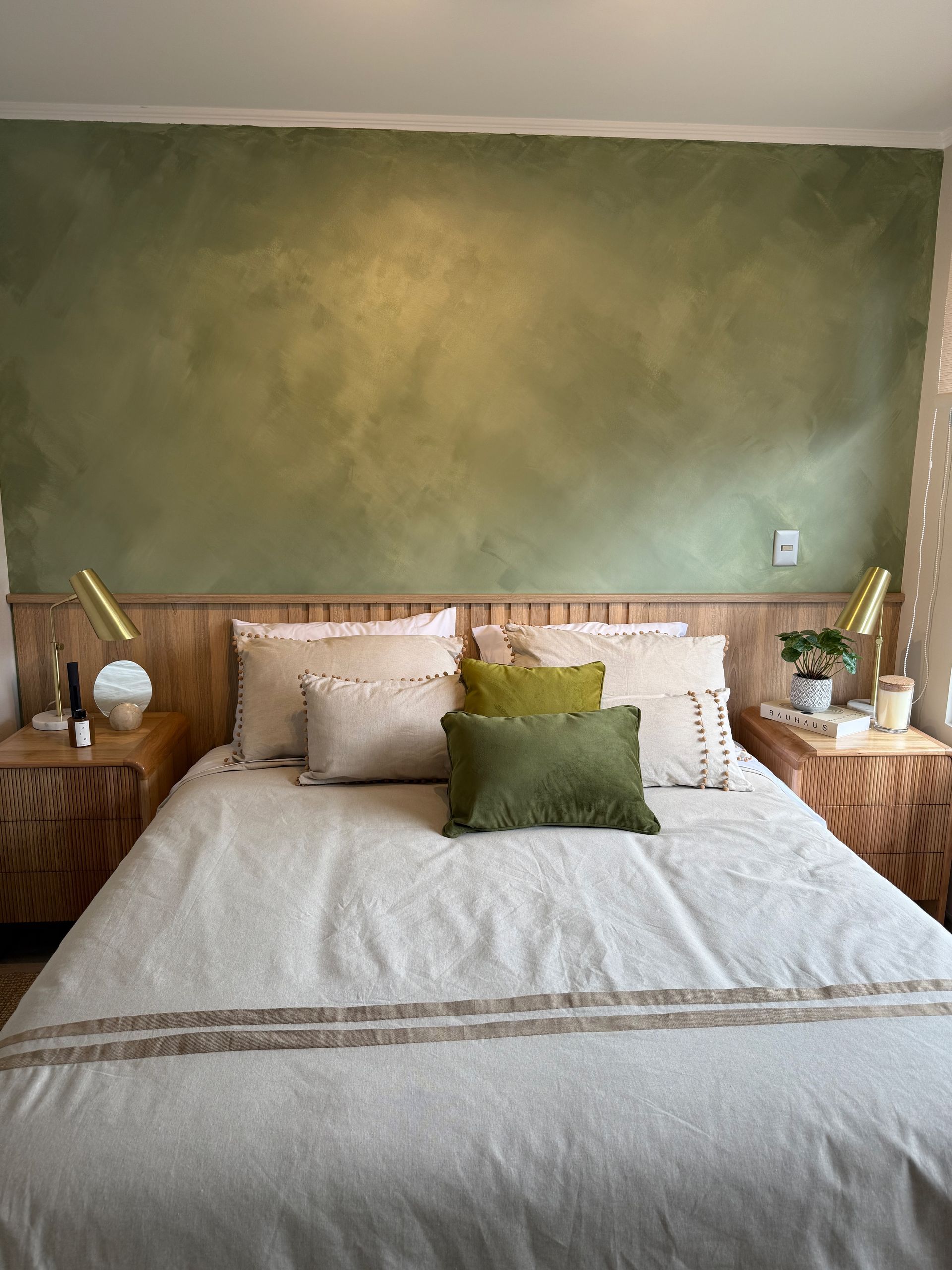 Cama bien hecha con ropa de cama blanca, almohadas beige y una pared decorativa verde en un dormitorio acogedor.