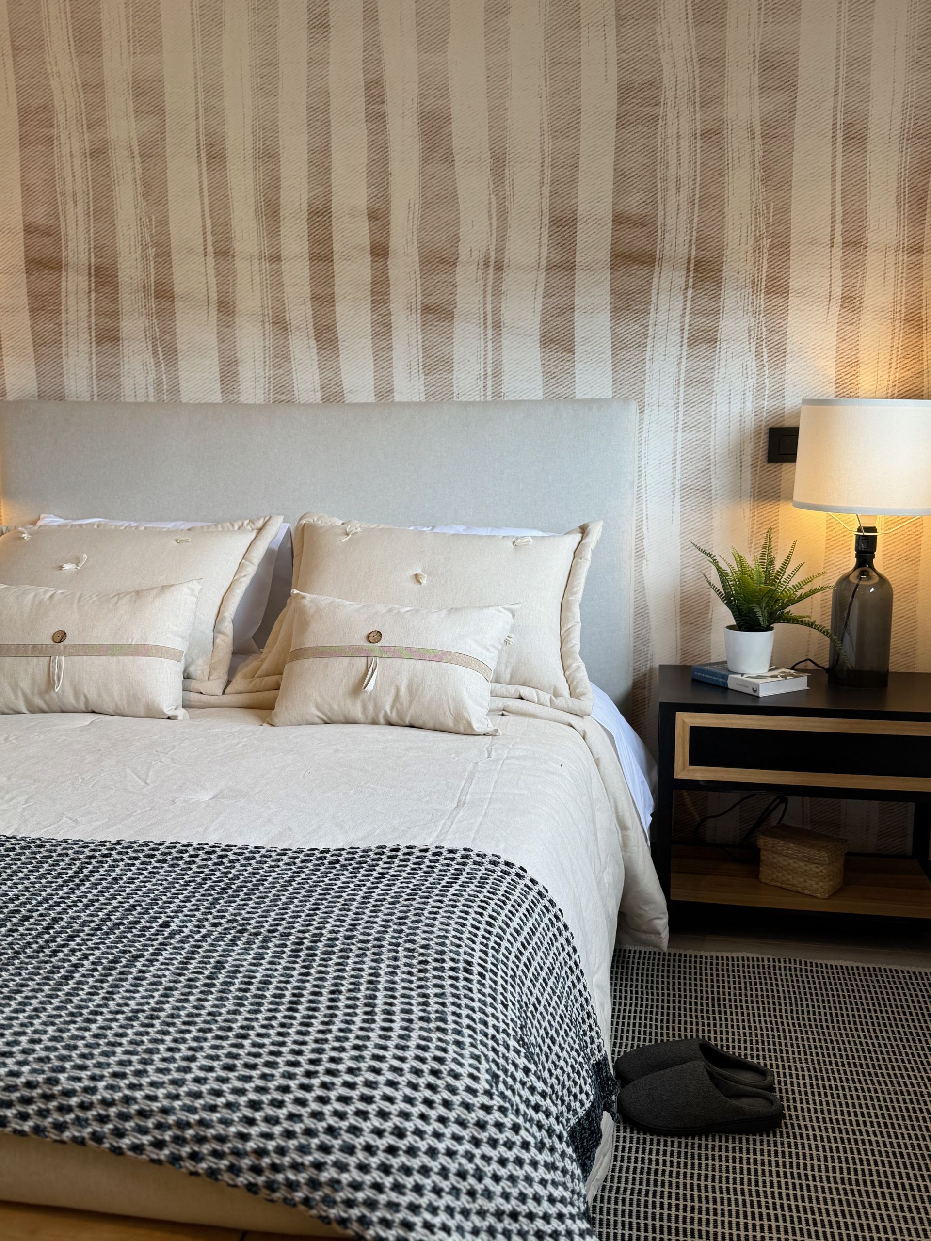 Habitación acogedora, una manta estampada, una lámpara de noche y una planta en maceta sobre la mesita de noche.