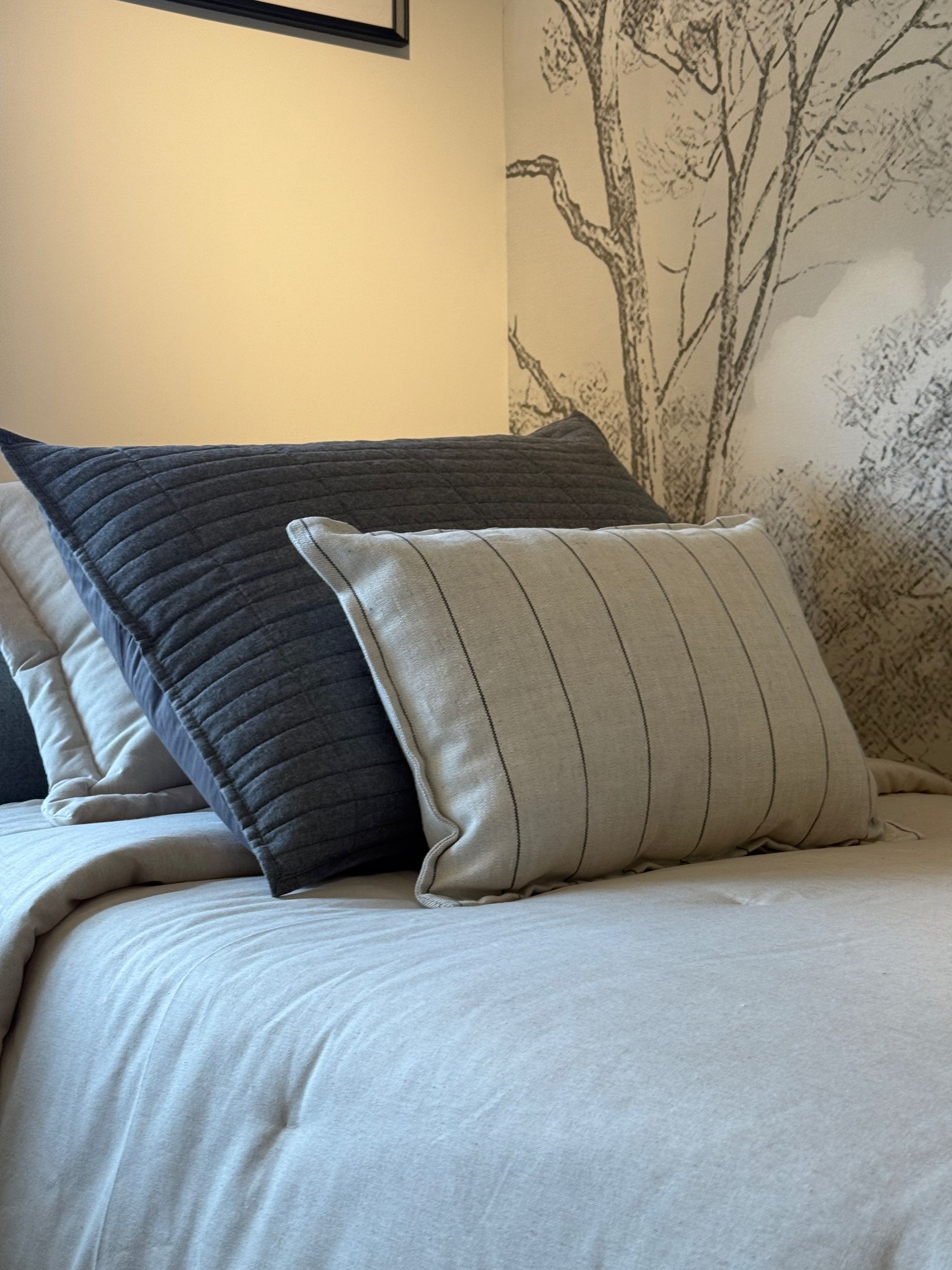 Cama bien hecha con sábanas blancas y almohadas grises superpuestas en un dormitorio con iluminación tenue.