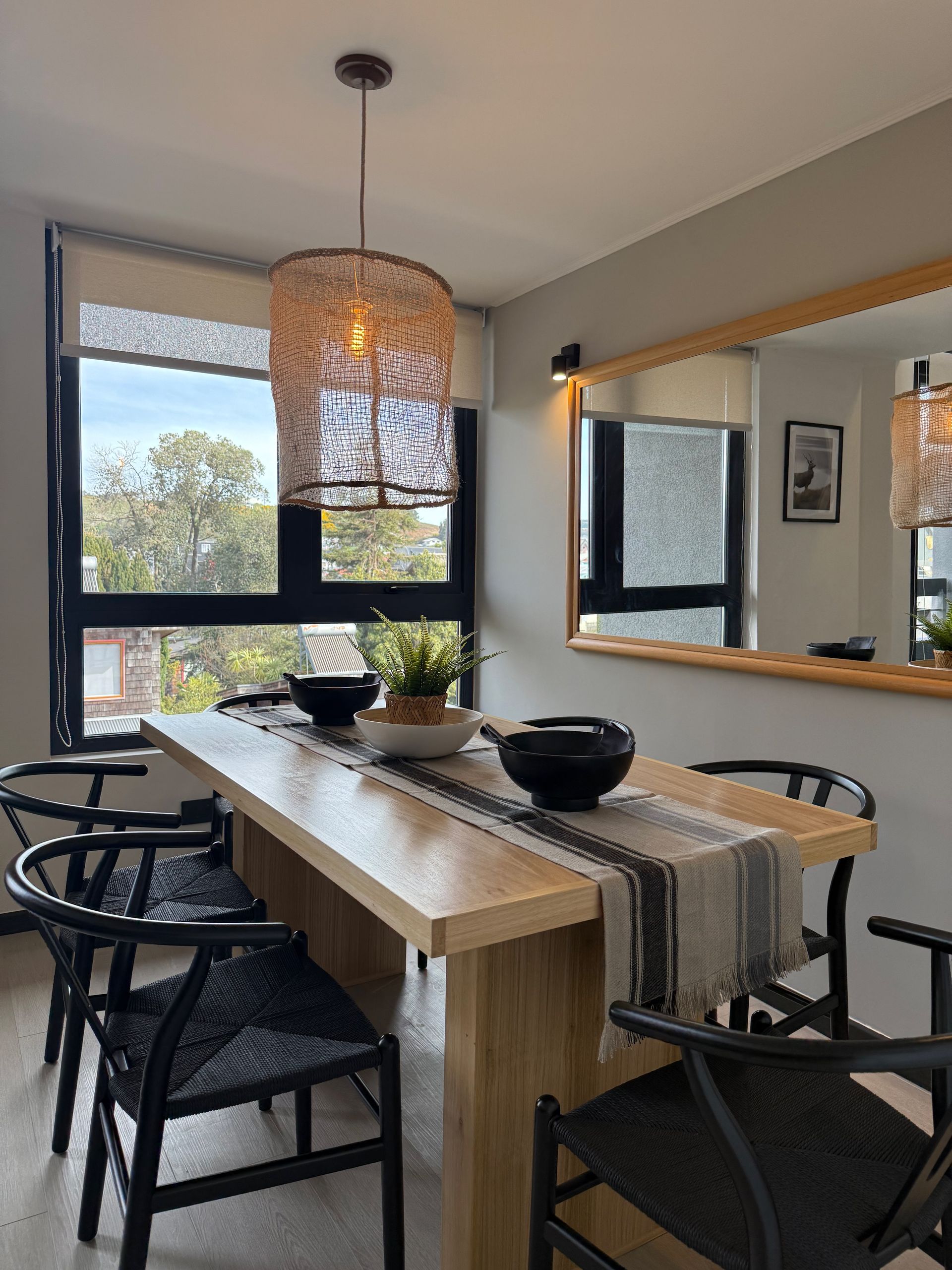 Comedor moderno con mesa de madera, sillas negras, lámpara colgante tejida y gran ventanal.
