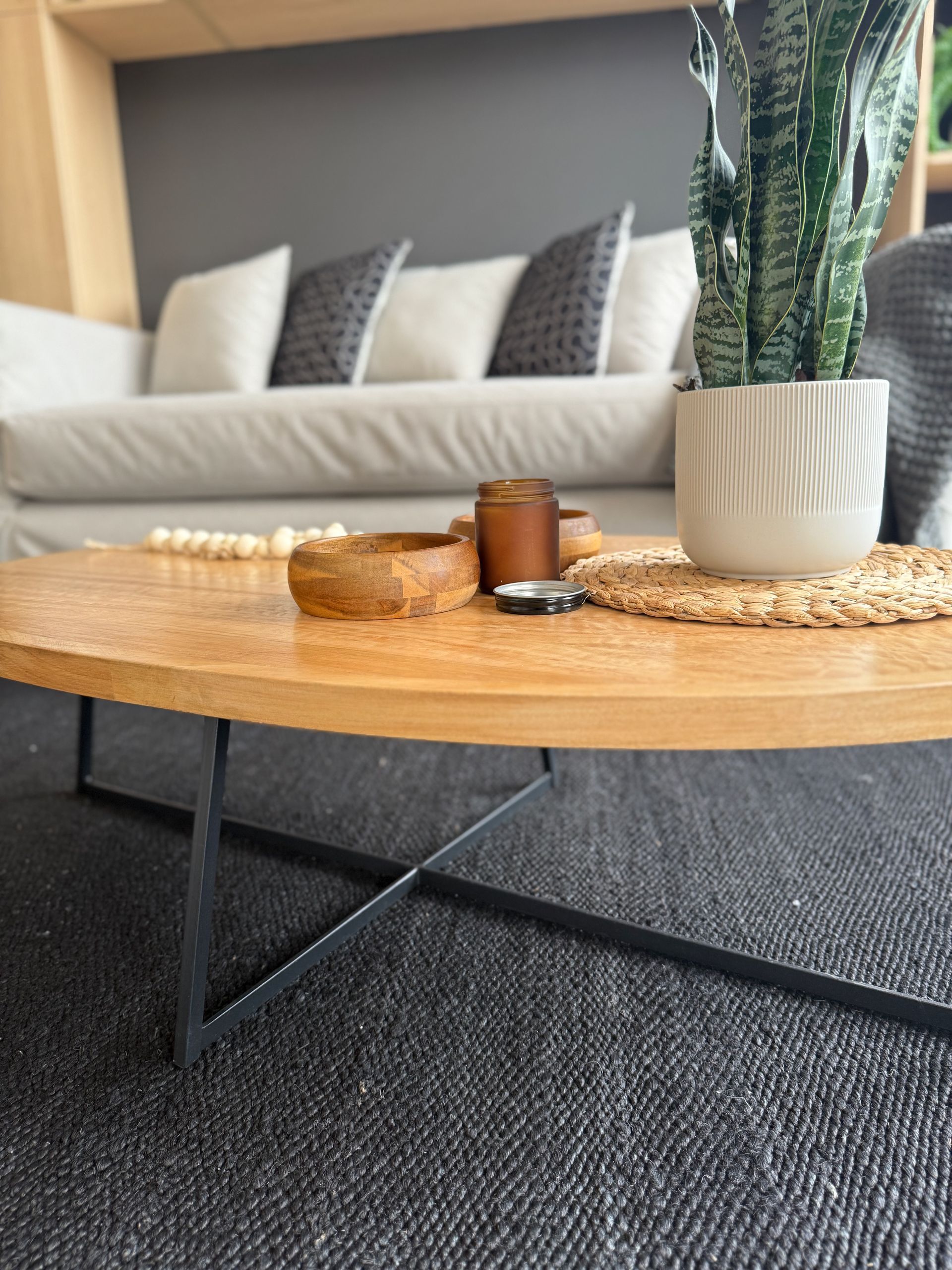 Sala de estar moderna con sofá gris, cojines estampados y una mesa de centro redonda de madera con una planta y velas.