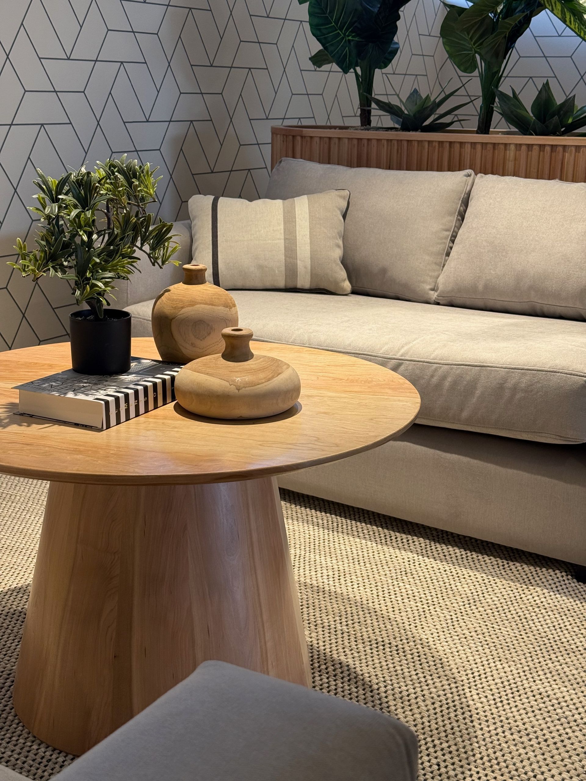 Acogedora sala de estar con sofá beige, mesa de centro redonda de madera, plantas y pared estampada.