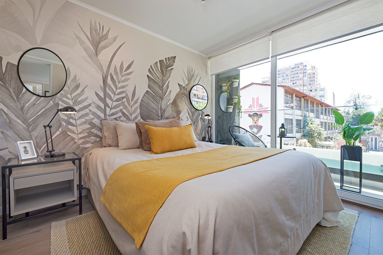 Habitación luminosa con cama, manta amarilla, mural de hojas en la pared.
