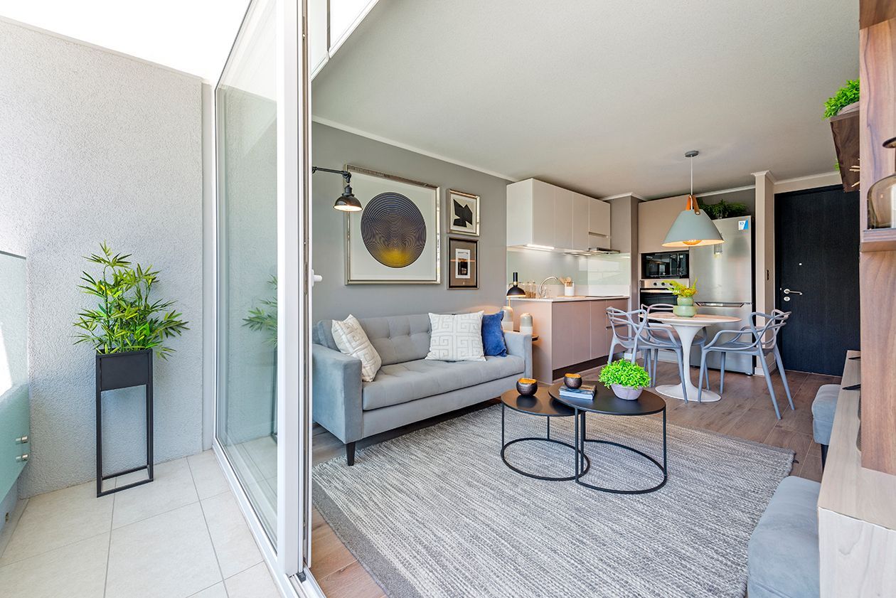Salón moderno de planta abierta con sofá gris, zona de comedor y plantas en el balcón bajo una brillante luz natural.